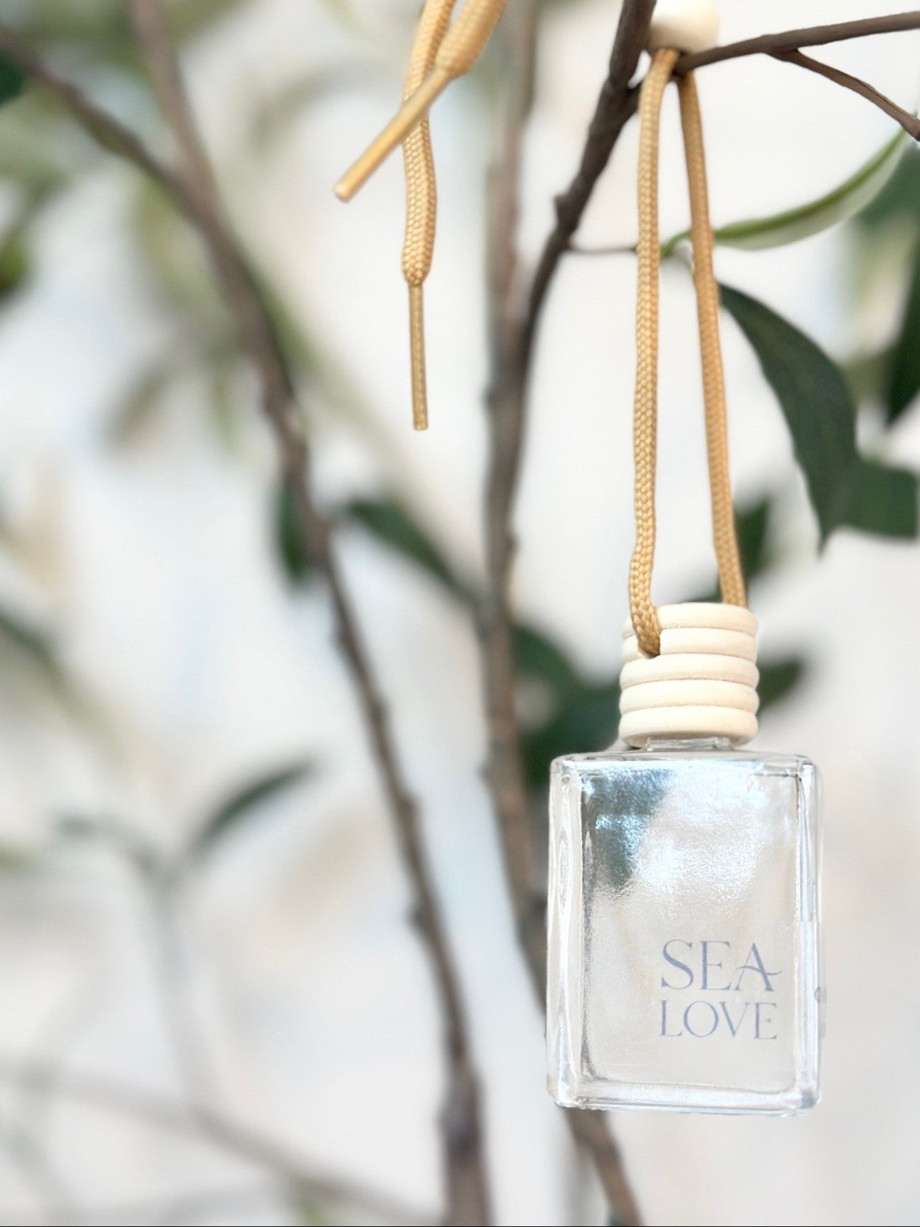 A translucent perfume bottle with the label "sea love" hanging from a beige ribbon on a green leafy branch, with a blurred background suggesting a serene and natural setting.