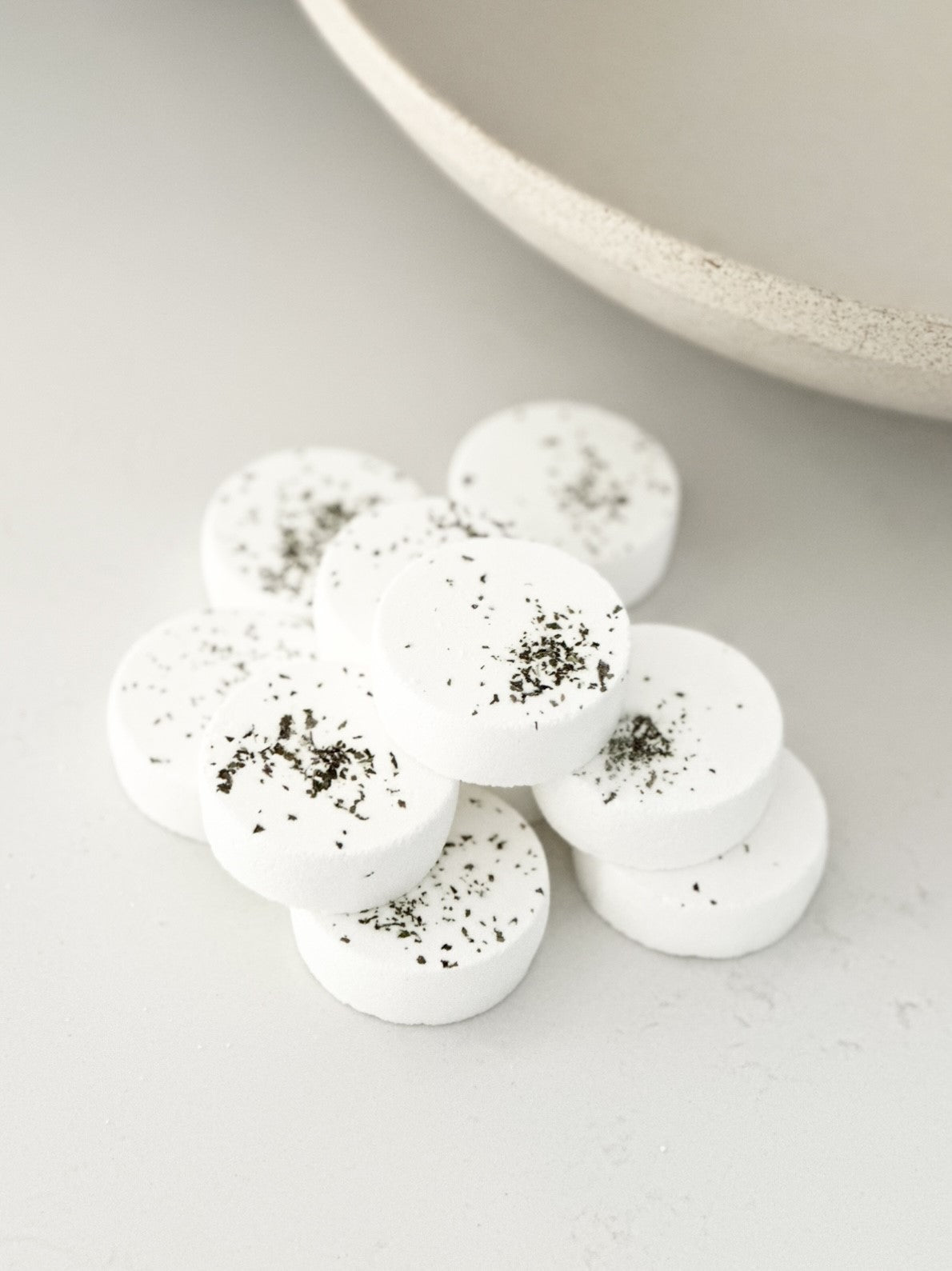 White, disc-shaped bath bomb with specks, arranged neatly on a light-colored surface next to a beige bowl.