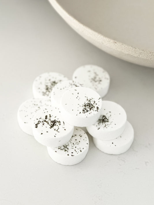 White, disc-shaped bath bomb with specks, arranged neatly on a light-colored surface next to a beige bowl.